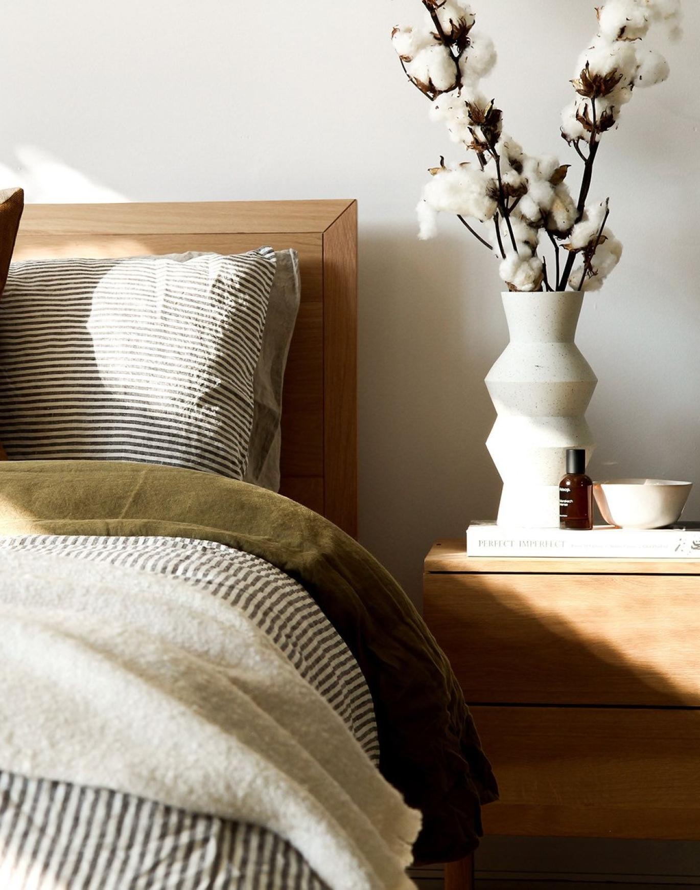 swath of sunlight shines across a well made bed and bedside table with a vase of cotton plants